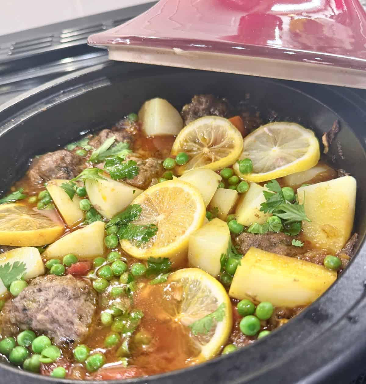 A red tagine top sits atop a tagine of meaballs with vegetables.