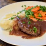 Salisbury Steak with Gravy on a white plate with mashed potatoes and peas and carrots.