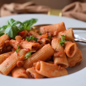 Wide fat rigatoni macaroni along with a rich tomato sauce in a white bowl with garnish.