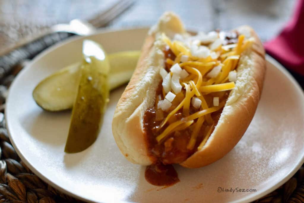 Plate with a dressed chili dog with onions and cheese being served with pickles.