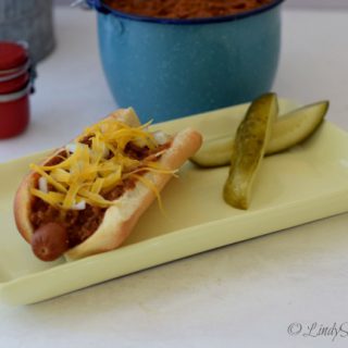 Lindy's Chili Gravy on a Chili Dog with pickles on a yellow plate.
