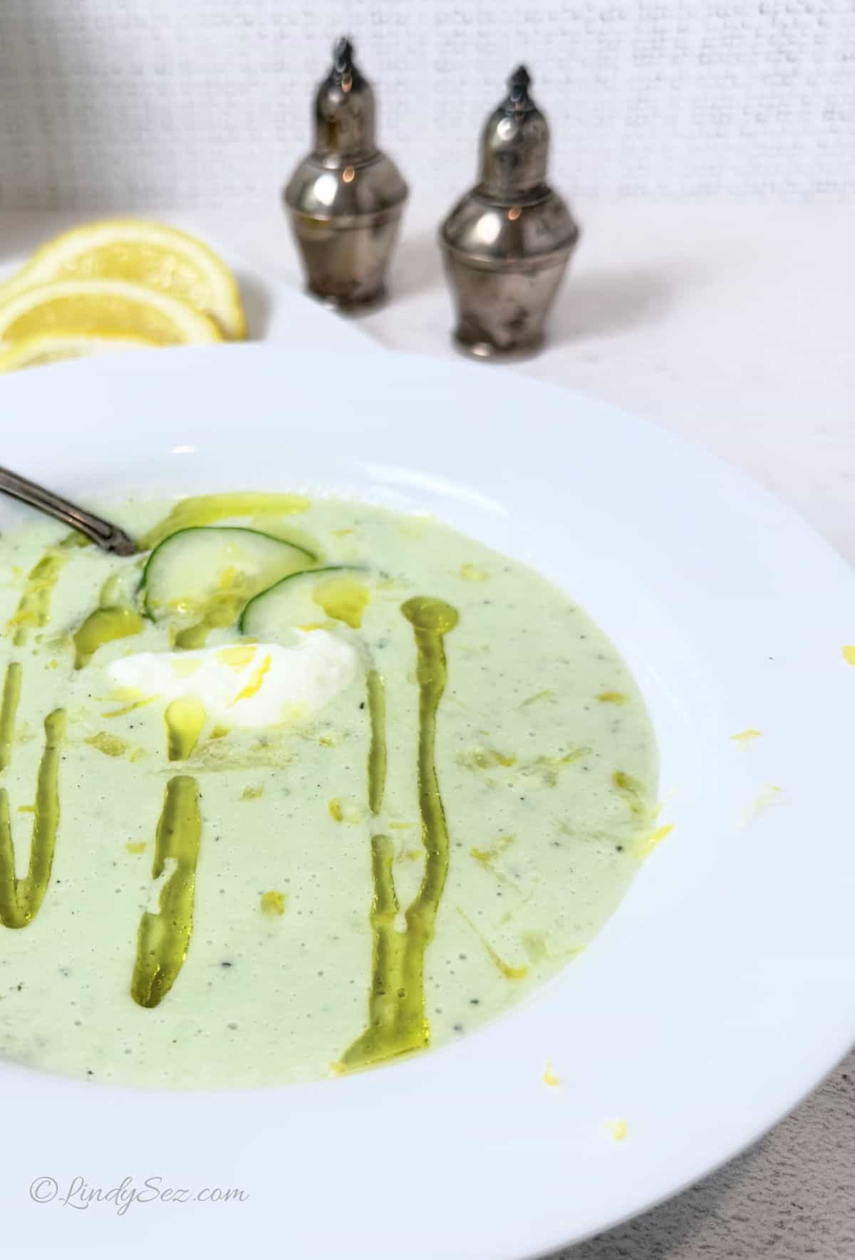 A white bowl of soup containing cucumber, yogurt, and llemon.
