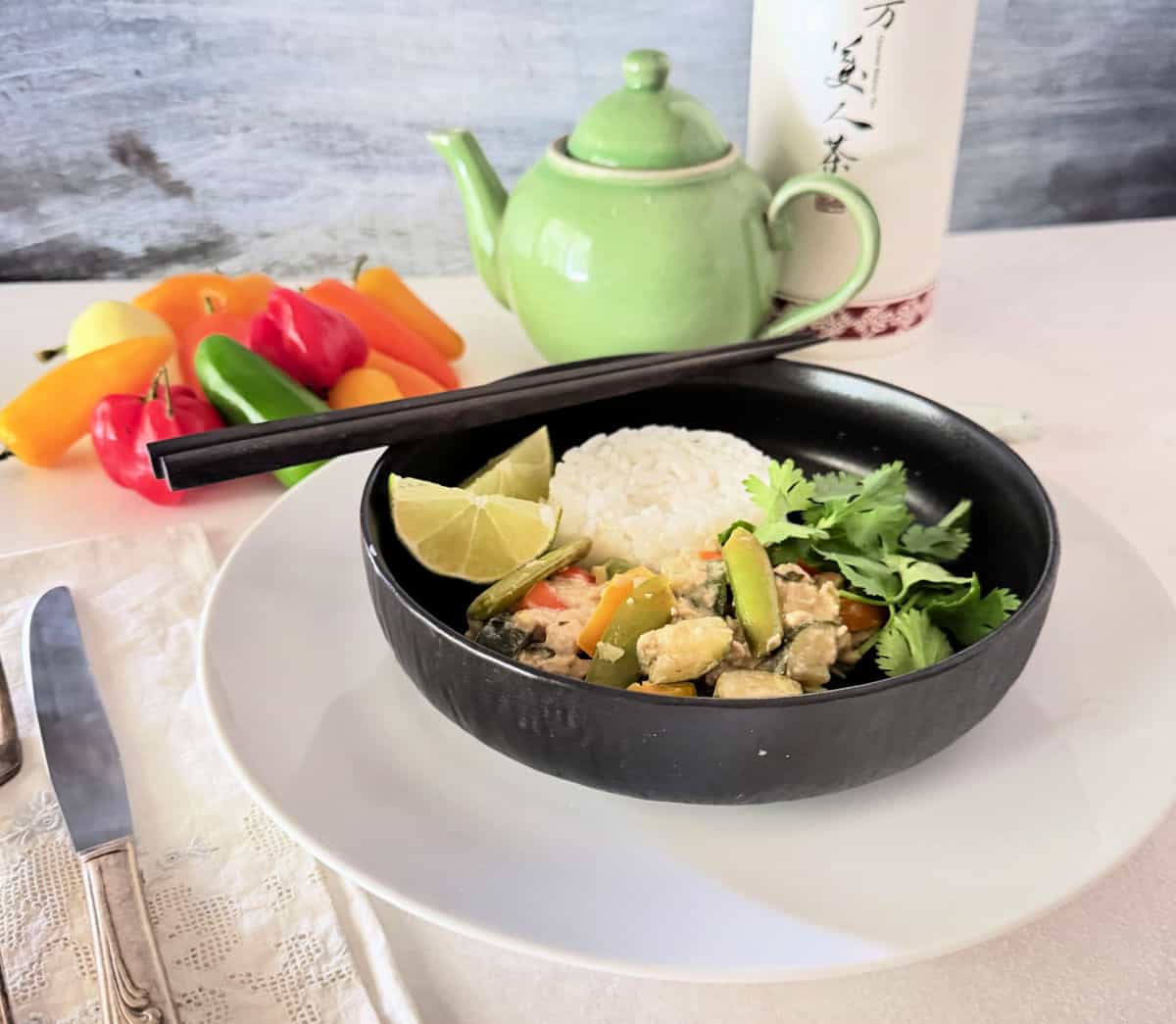 A black bowl with Thai Stir Fry along with peppers and a green ceramic tea pot.