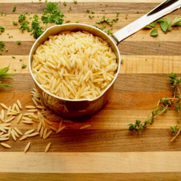 Raw orzo in a measuring cup with herbs scatted around.