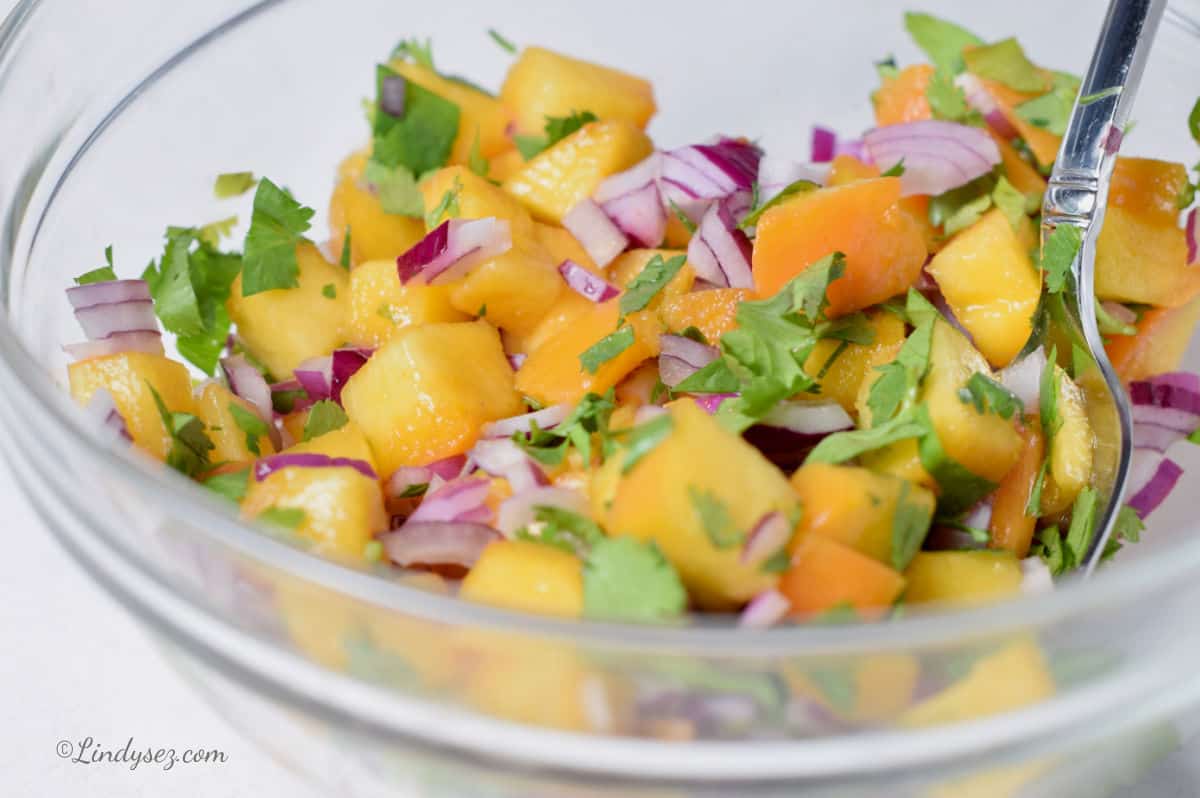 A tasty peach salsa in a glass bowl with cilantro, red onion, and lime juice.
