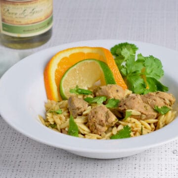 An orange and lime slice enhance a white bowl with orzo and pork cubes.
