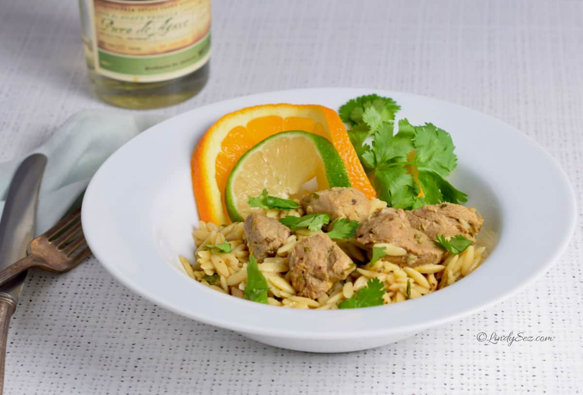 An orange and lime slice enhance a white bowl with orzo and pork cubes.