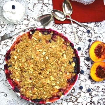 A blueberry and peach crumble on a lace tablecloth with peaches and whipped cream beside the dish.