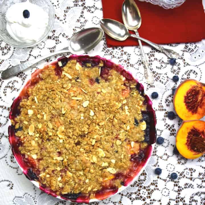 A blueberry and peach crumble on a lace tablecloth with peaches and whipped cream beside the dish.