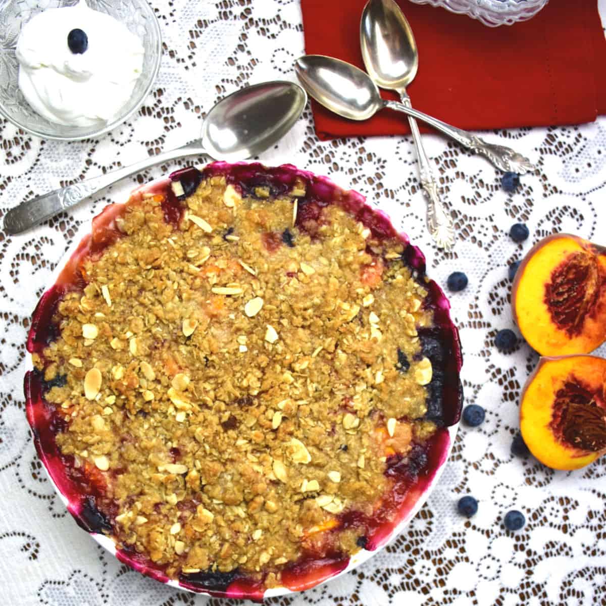 A blueberry and peach crumble on a lace tablecloth with peaches and whipped cream beside the dish.