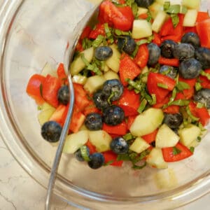 A glass bowl with colorful tomatoes, blueberries, and cucumber with fresh basil.