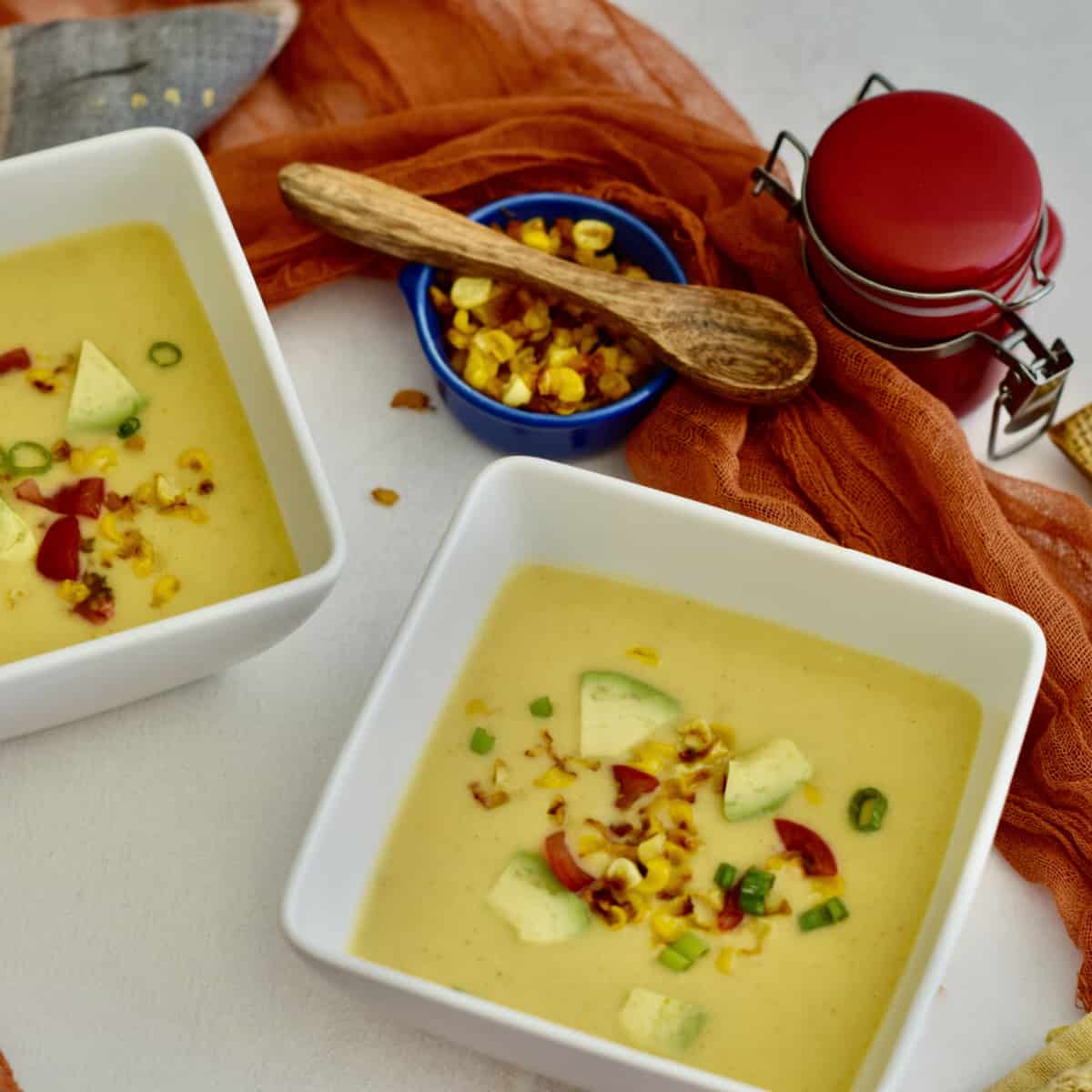 Two square white bowls with corn soup and toppings on a table with roasted corn and a wooden spoon.