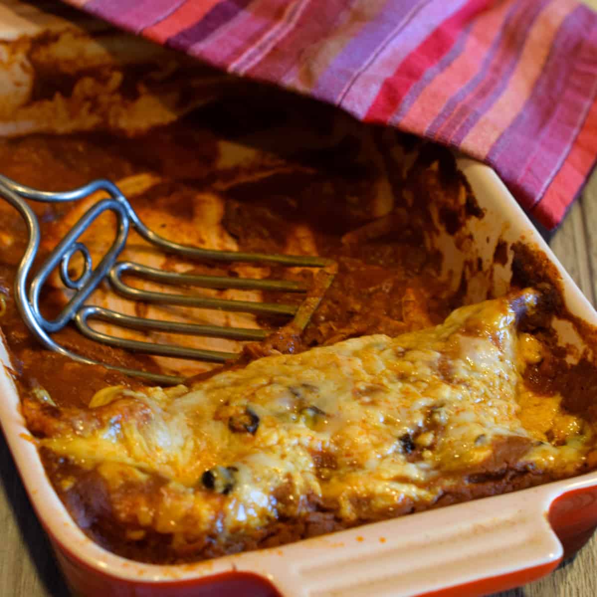 A shredded chicken enchilada in a baking dish with a striped napkin and old fashioned  serving spoon.