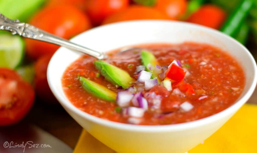 A chunky bowl of Gazpacho with fresh vegetables in the background.