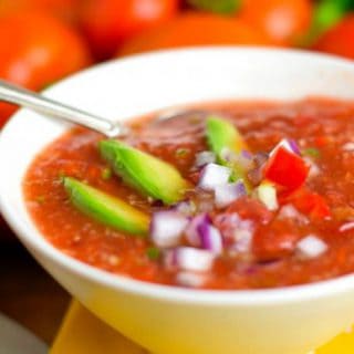 A chunky bowl of Gazpacho with fresh vegetables in the background.
