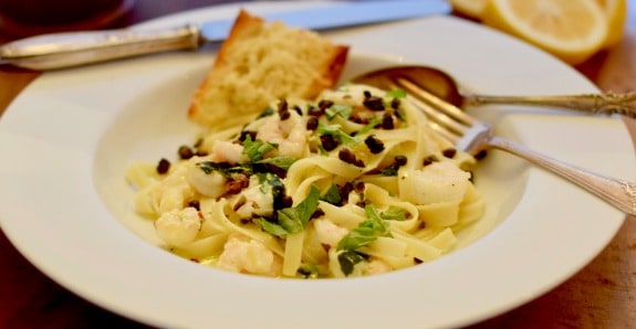 A white bowl with lemony pasta tossed with shrimp and topped with fried capers