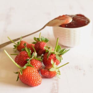 Fresh strawberries scattered with a small bowl of Easy Fresh Strawberry Jam with Balsamic and a spoon.