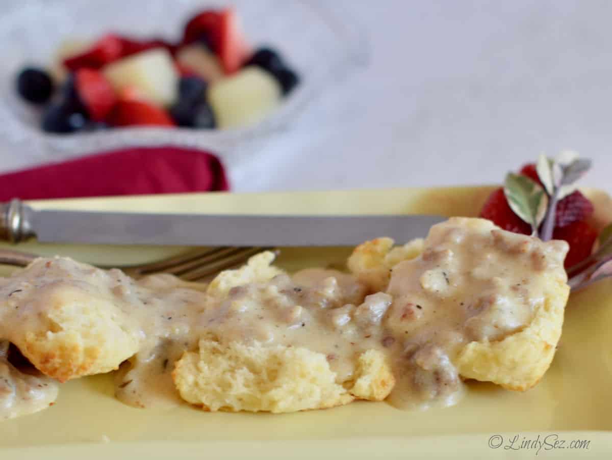 Biscuits and gravy on a yellow plate with fruit in the background.