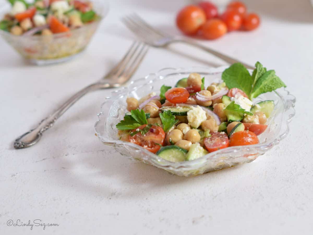 Tomatoes, garbanzo beans, feta cheese, fresh herbs, combine to make the colorful salad that is being served in an ornate glass bowl.
