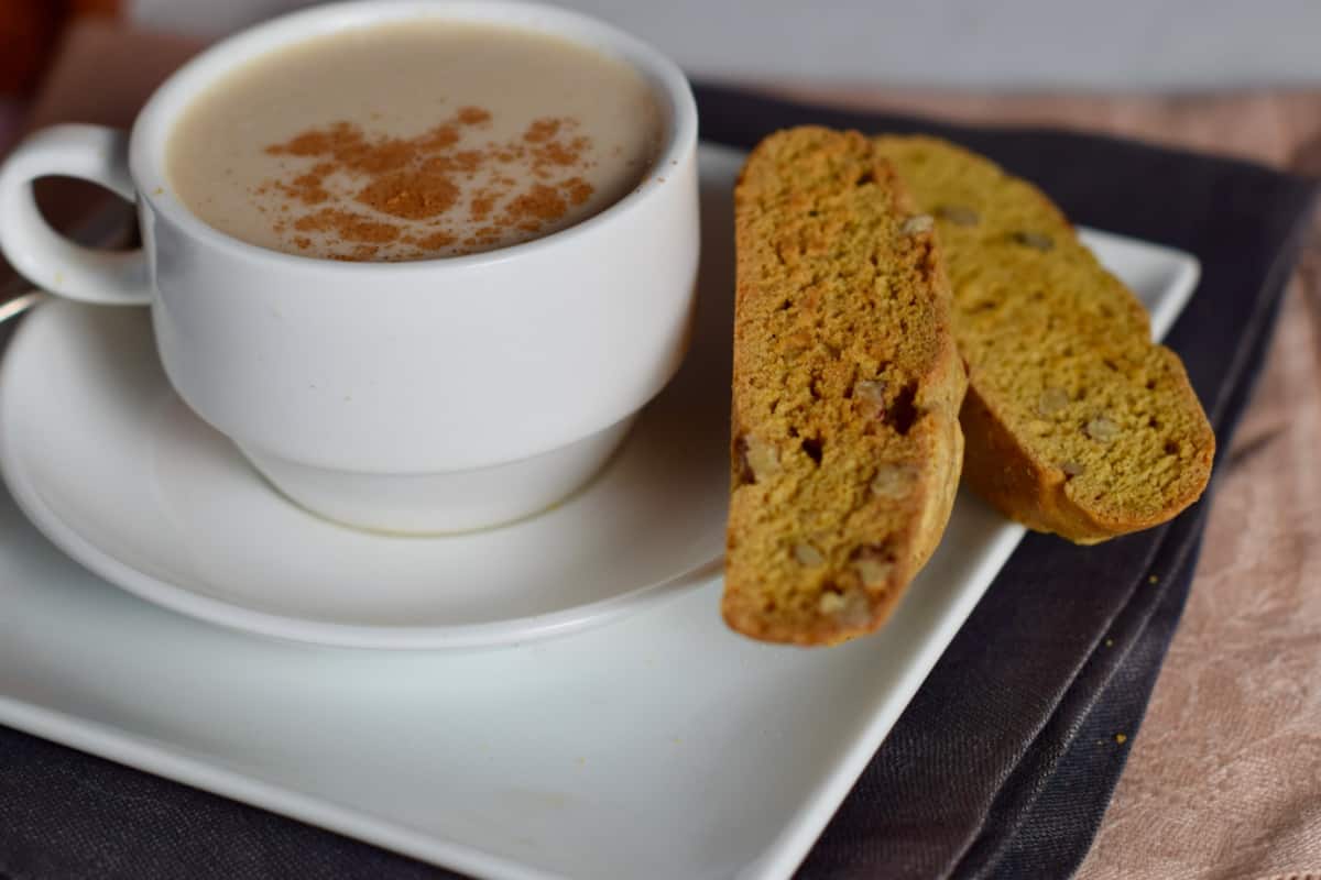 A cup of coffee with two pumpkin rum biscotti with pecans on a square white plate.